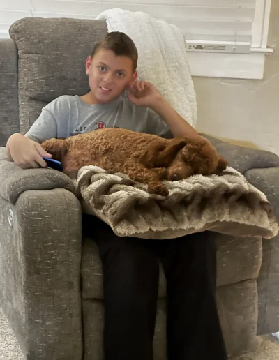 A boy sits on a gray recliner holding a remote, with a small brown mini poodle puppy lying on a fuzzy beige blanket on his lap. The boy is smiling and has one hand resting on his ear.