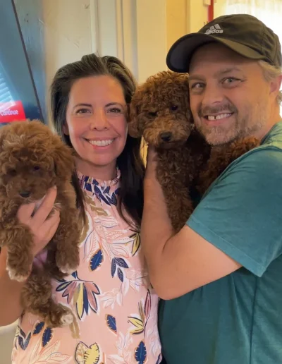 A smiling woman and man stand close together indoors, each holding adorable mini poodle puppies. The woman wears a light patterned blouse, and the man wears a green shirt and cap. A television is in the background. Perfect day with poodle puppies Utah!.