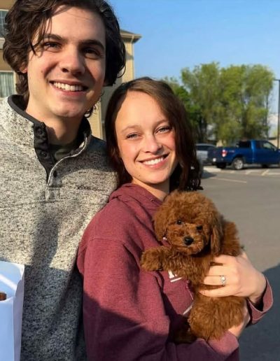 A smiling couple stands outdoors, the woman cradling a small, fluffy brown puppy in her arms. If you love mini poodle puppies, you'll adore this sweet scene set against cars and trees under a clear sky.