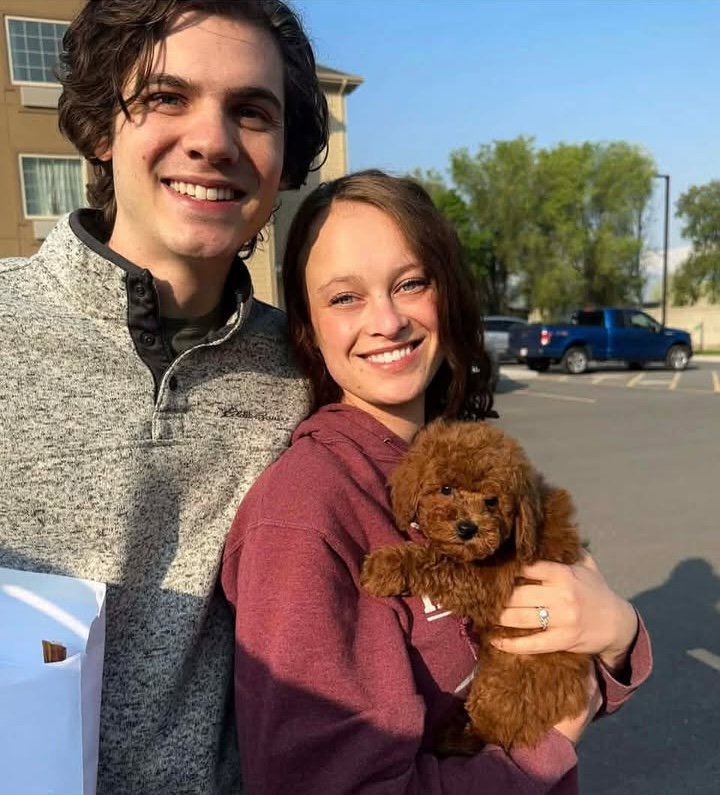 Mountain Valley Poodles - Stephanie Sanders - Mini Poodle Puppies For Sale in Utah A smiling couple stands outdoors, the woman cradling a small, fluffy brown puppy in her arms. If you love mini poodle puppies, you'll adore this sweet scene set against cars and trees under a clear sky.