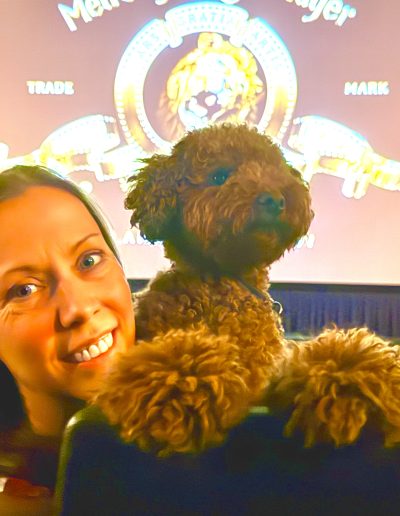 A smiling woman and a fluffy brown mini red poodle sit in a movie theater with the MGM logo displayed on the screen in the background.