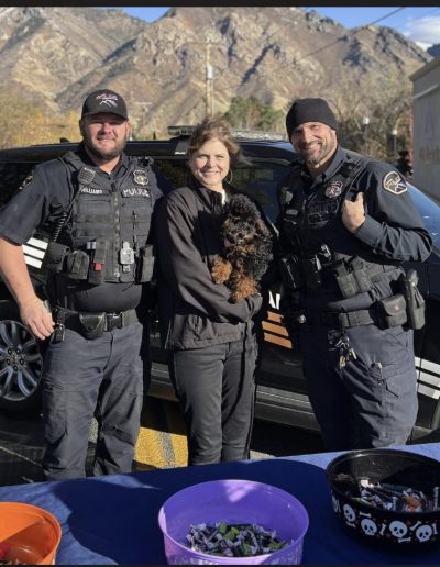 Three smiling police officers stand outside in front of a police car and mountains, with one person in the middle holding an adorable mini red poodle. Bowls of candy sit on a table in the foreground, perfect for poodle puppies Utah fans to enjoy.